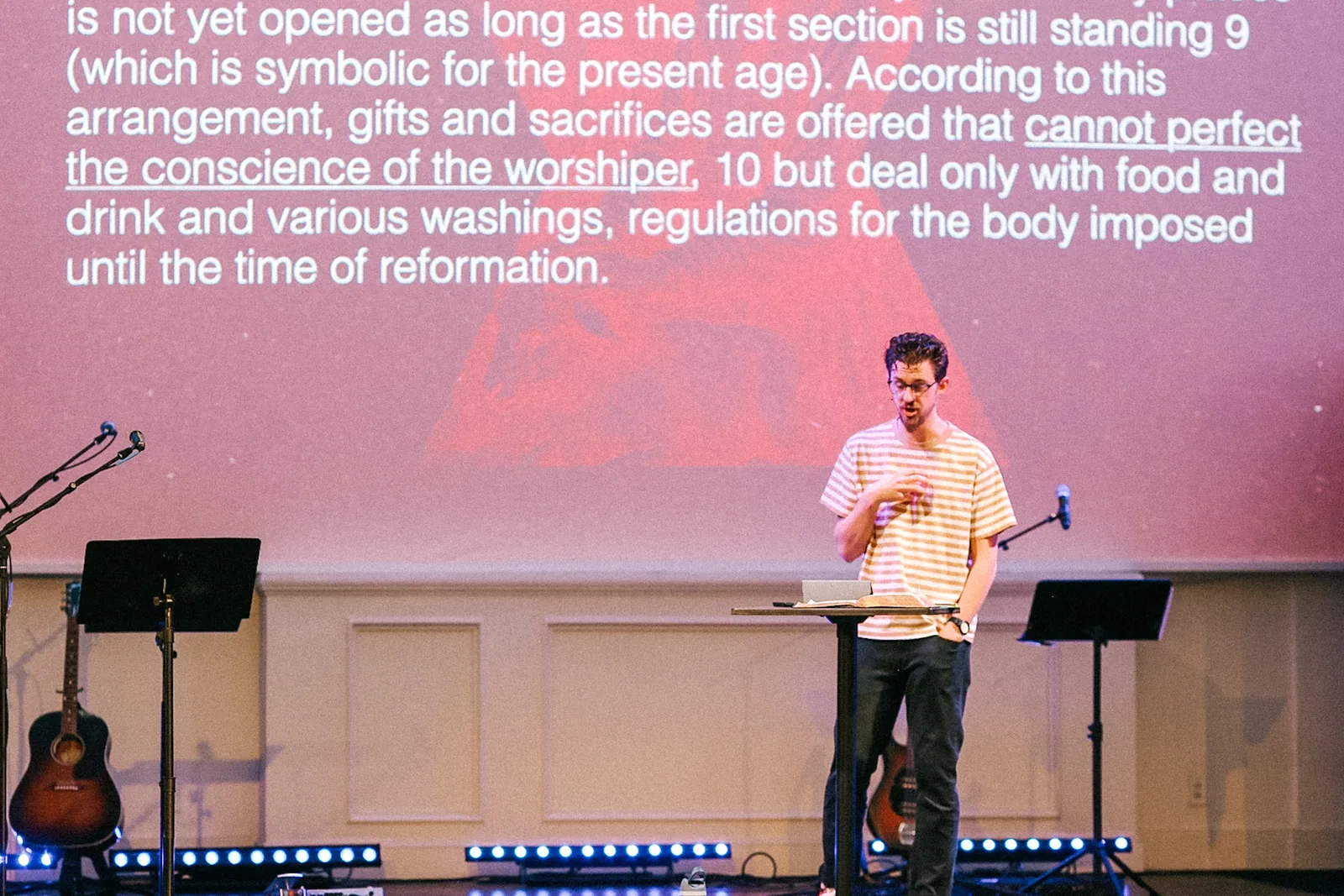 A younger pastor teaching at a lectern with a passage from Hebrews projected behind him