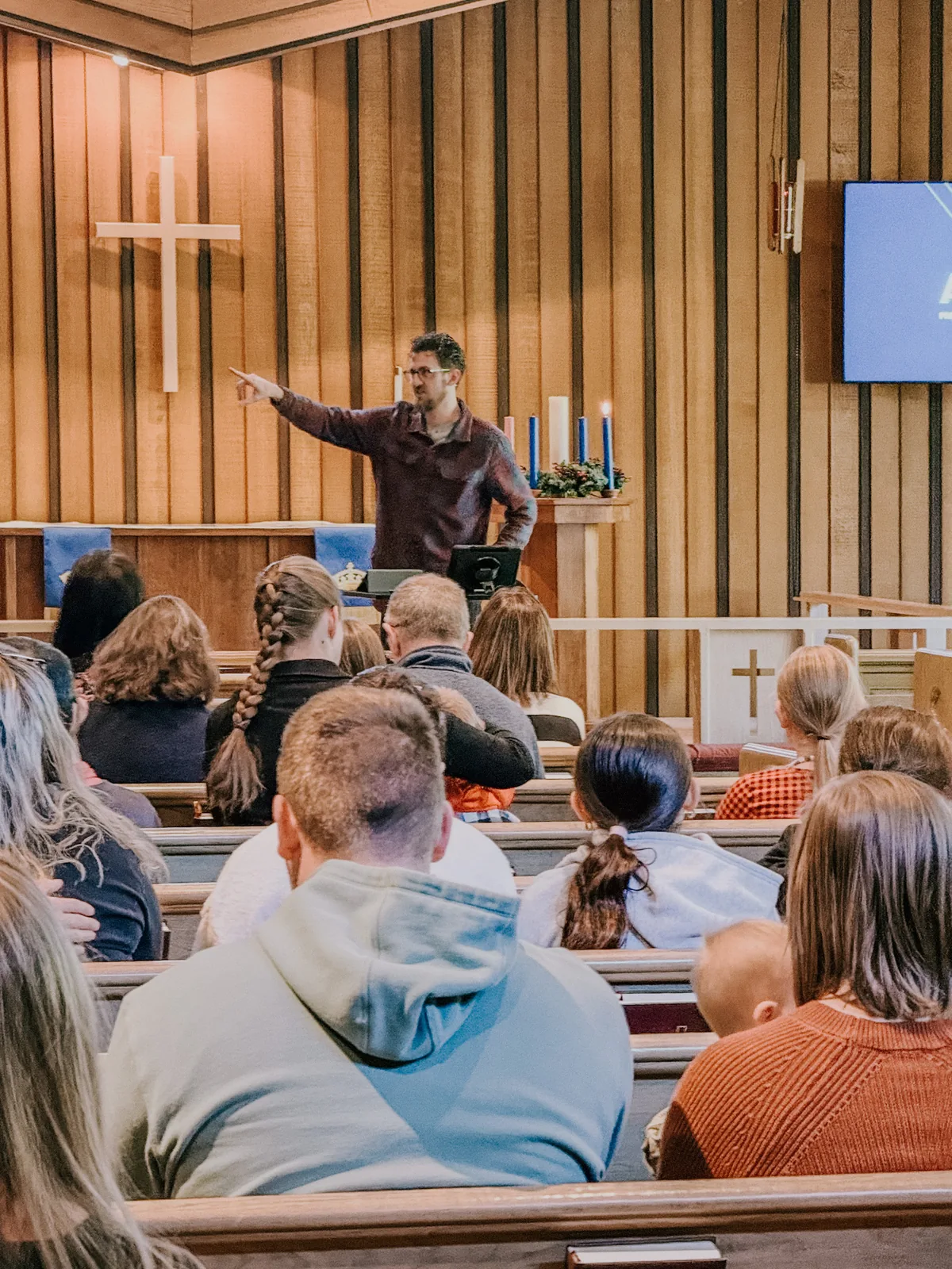 A younger pastor pointing toward his seated congregation from the front of a wood-panelled sanctuary